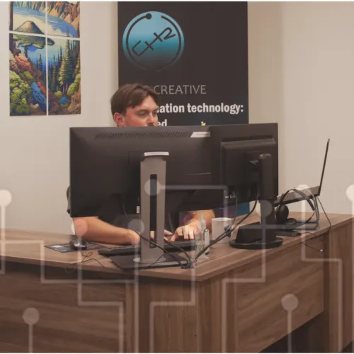 Two men working at separate desks in a modern office with computer monitors and large windows.