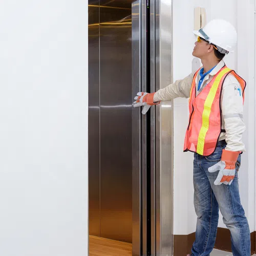 Construction worker wearing safety gear opening elevator doors indoors, standing against white walls.