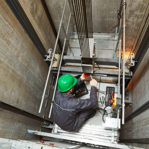 Technician in green helmet repairing elevator mechanism inside a concrete elevator shaft looking downward