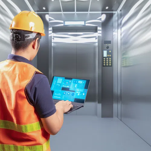 Engineer in safety gear using a laptop with digital data inside a modern stainless steel elevator.