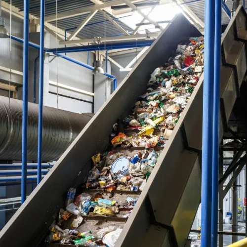 Conveyor belt carrying mixed plastic waste inside a recycling facility with industrial pipes and metal structures.