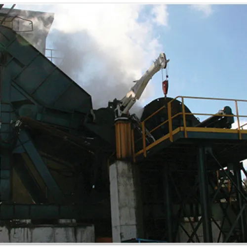 Industrial machine emitting smoke at a factory under blue sky with scattered clouds and surrounding trees.