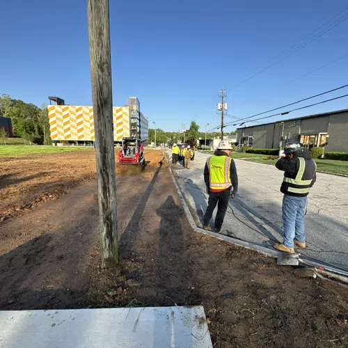 Construction workers in safety gear working on roadside excavation near utility poles under clear blue sky.