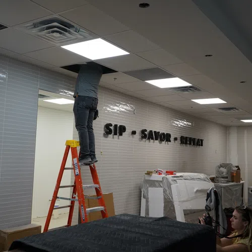Worker on ladder installing ceiling tiles in a room with tiled walls displaying 'SIP SAVOR RELAX' sign.