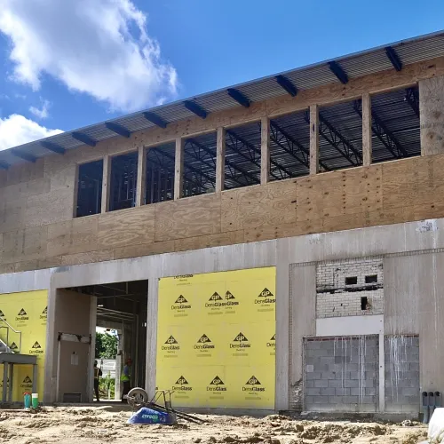 Partially constructed two-story building with plywood upper walls, concrete lower walls, and metal staircase under blue sky.