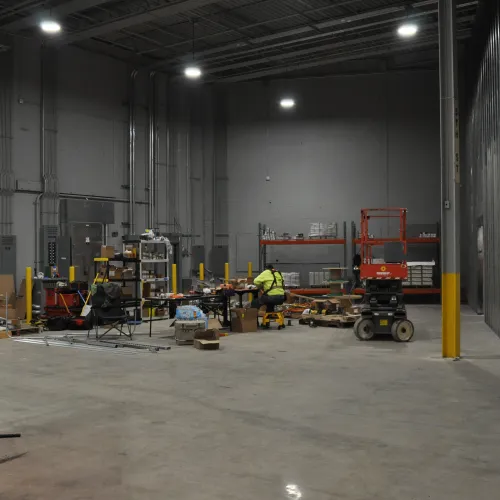 Spacious industrial warehouse interior with construction equipment, workers, and storage shelves under bright lighting.