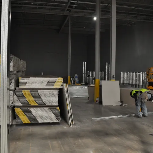 Warehouse interior with stacked drywall sheets and a worker installing drywall on the floor under construction.