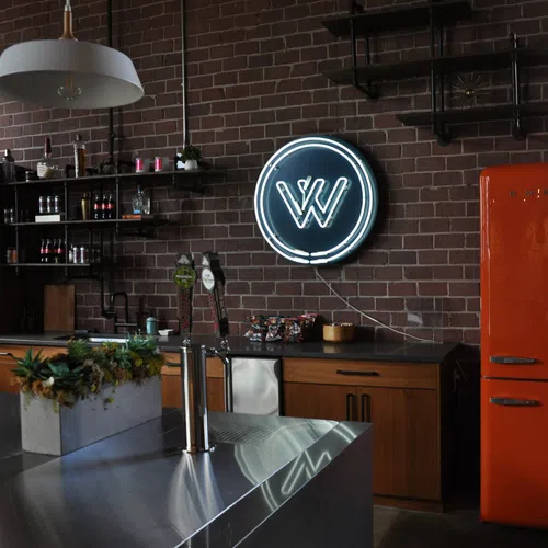 Industrial style kitchen with brick walls, neon sign, orange retro fridge, wooden cabinets, and modern stainless steel island.