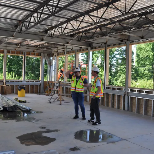 Two construction workers in safety gear discuss plans inside a metal-framed building under construction with large windows.