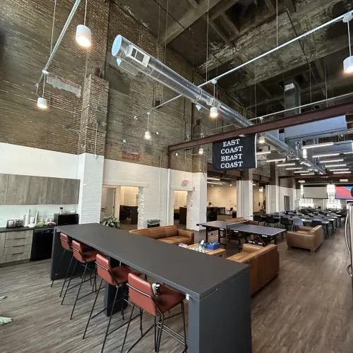 Spacious industrial-style office with high ceilings, lounge areas, kitchen, and multiple seating options under exposed ductwork.