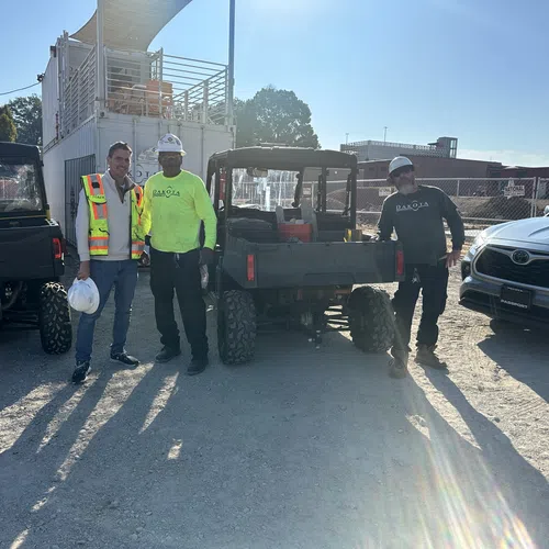 Three construction workers standing on a gravel site with utility vehicles and a silver car under bright sunlight.