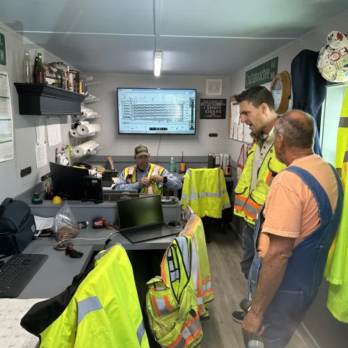 Construction workers in safety vests and gear inside an office discussing plans with computers and charts around.