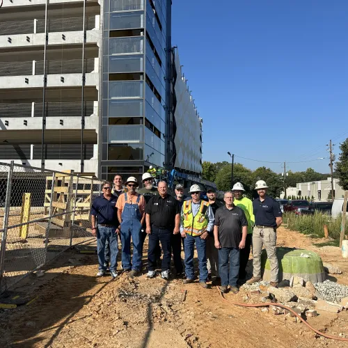 Group of construction workers posing at an active building site with a partially completed parking structure behind them.