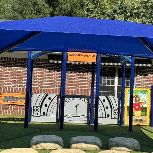 Outdoor children's play theater area with blue canopy, stage backdrop, and artificial grass seating resembling rocks.