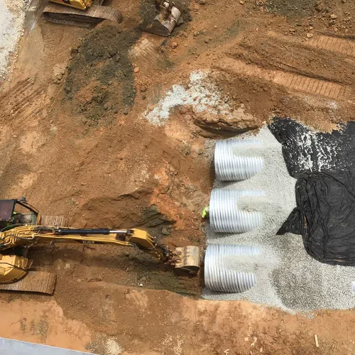 Construction site with excavator digging soil near large corrugated pipes and gravel bedding.