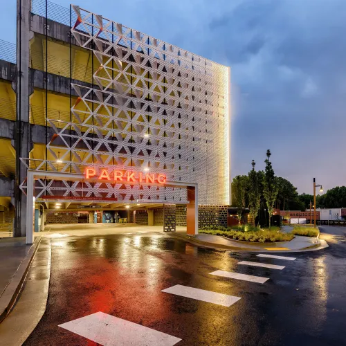 Modern parking garage entrance with illuminated geometric facade and wet reflective pavement at dusk.