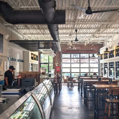 Modern cafe interior with high ceilings, wooden tables and chairs, glass display case, and menu boards.