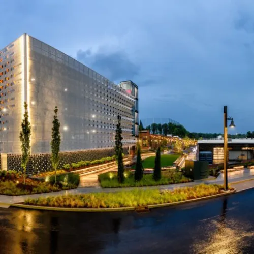 Modern illuminated parking garage and commercial buildings at dusk with wet roads and landscaped greenery.