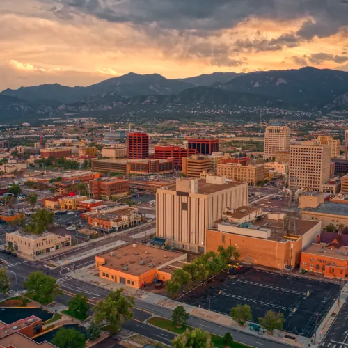 Aerial view of a city with diverse buildings under a cloudy sunset sky and mountains in the background.