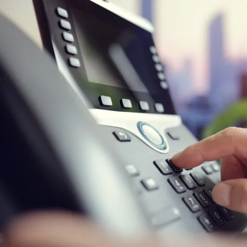 Close-up of a hand dialing numbers on a modern office IP phone with blurred city background.