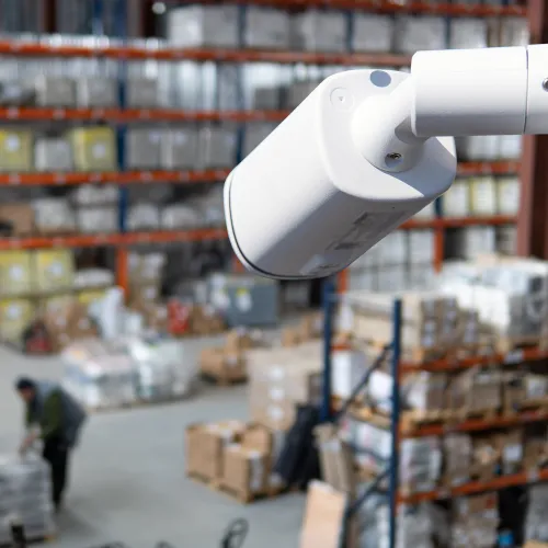 White security camera monitoring a large warehouse with shelves and workers moving packages.