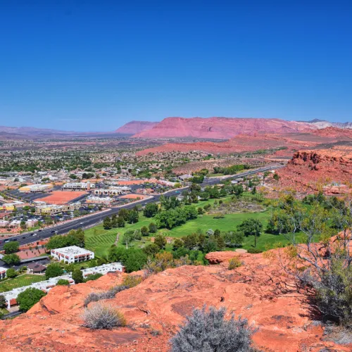 Aerial view of a town with red rock formations, green fields, and a clear blue sky in a desert landscape.