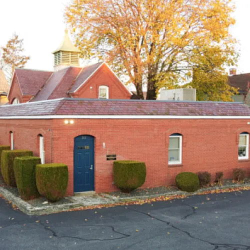Small red brick building with blue door, trimmed bushes, and large autumn tree behind it under clear sky.