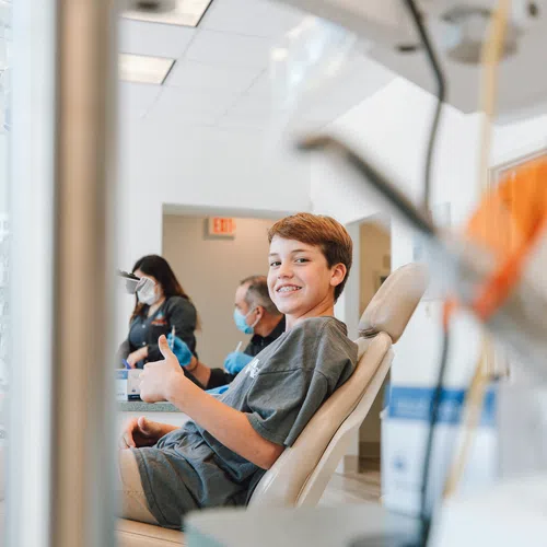 Teen boy smiling with braces, giving a thumbs up while sitting in a dental chair during a checkup.