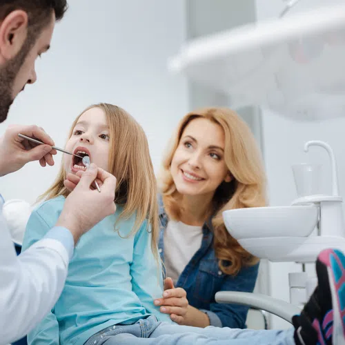 Dentist examining young girl's teeth while her mother watches supportively in dental clinic.