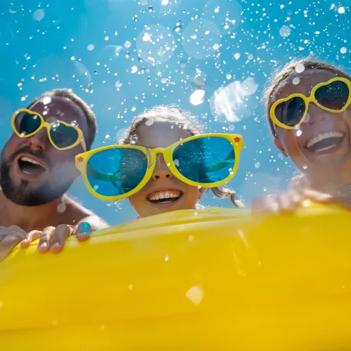 Family wearing oversized sunglasses and smiling while leaning on a bright yellow inflatable float in a sunny pool.