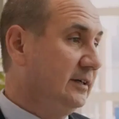 Close-up of a middle-aged man in a suit and red tie speaking, with a window background providing natural light.