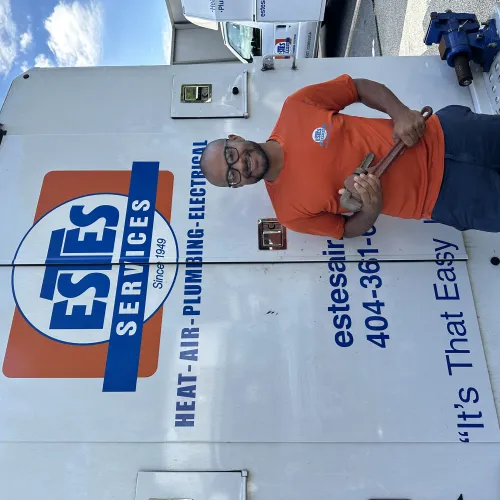 Estes Services Plumber  in orange shirt holding a pipe wrench standing next to an Estes Services branded truck with service information.