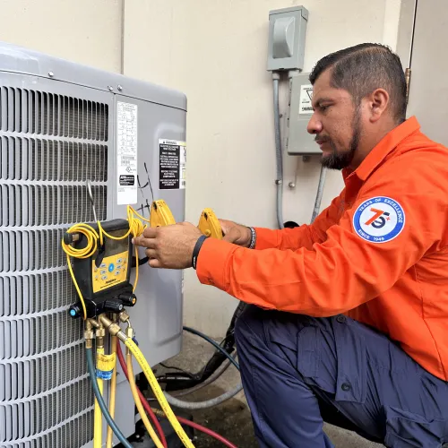 Estes Services Technician in orange uniform repairing and testing an HVAC air conditioning unit with digital gauges and wires outdoors.