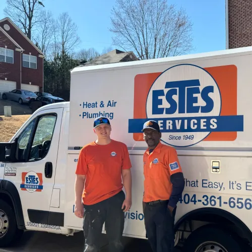 Two Estes Services plumbers in orange uniforms stand beside a company service van in a residential neighborhood.