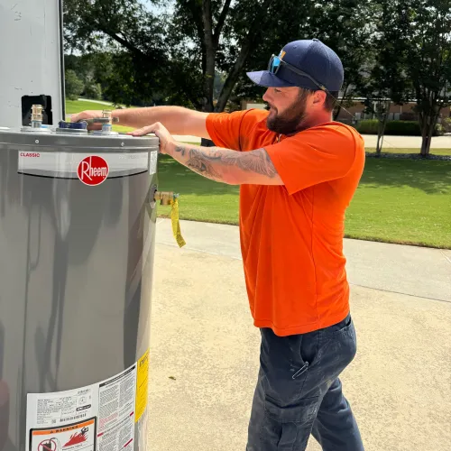 Estes Services Plumber in orange shirt installing or inspecting a Rheem water heater outdoors on a sunny day.