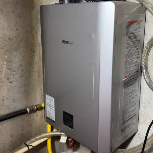 Silver Rinnai tankless water heater installed on a concrete wall with connected pipes and a white bucket below.