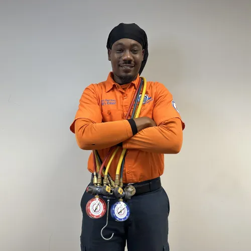 Technician in orange shirt with HVAC gauges and hoses standing confidently against a plain wall.