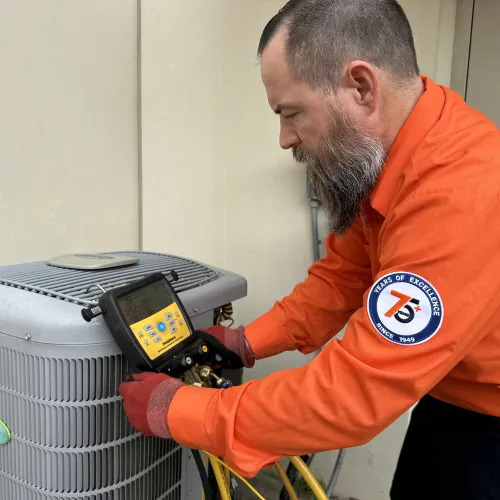 Technician in orange shirt using diagnostic tools on a Carrier air conditioning unit outdoors against beige wall.