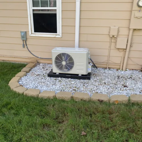 Outdoor HVAC unit installed on gravel bed bordered by bricks beside beige house with grass foreground.