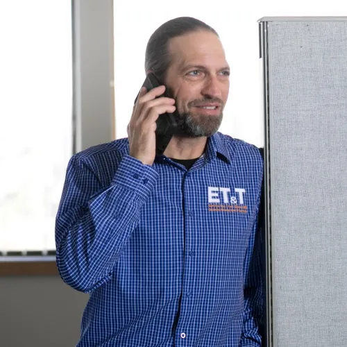 Man in blue checkered shirt talking on phone in bright office with large windows and gray partition.
