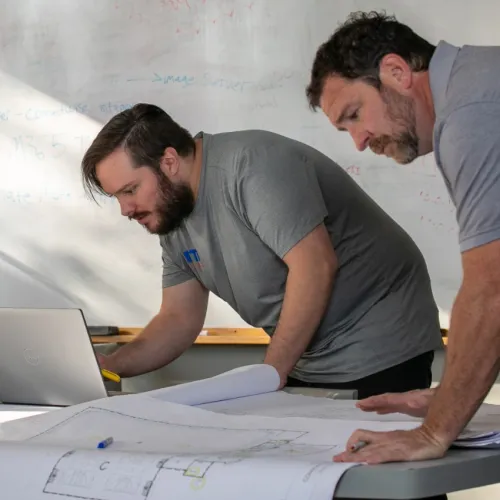 Three professionals collaborate over architectural plans and a laptop in a bright, modern office meeting room.