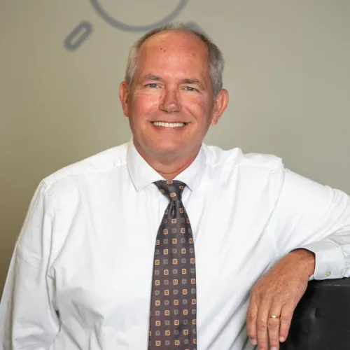 Smiling middle-aged man in a white shirt and patterned tie leaning on a black surface with light bulb drawings on wall.