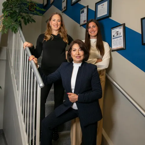 Three professional women posing on an indoor staircase with framed certificates on the wall and a plant nearby.