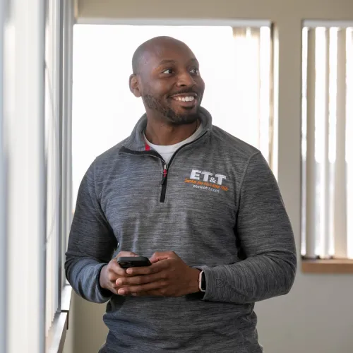 Smiling man in gray ET&T sweatshirt holding phone near a window with blinds in a bright room.