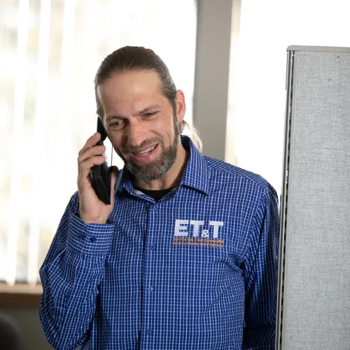 Man in blue checkered shirt talking on phone in a bright office space with cubicle divider.