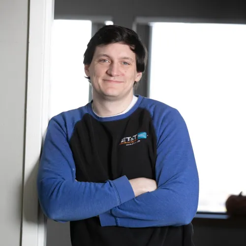 Young man with short dark hair, wearing a black and blue long sleeve shirt, standing with arms crossed indoors.