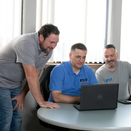 Four men in casual workwear gathered around a laptop in a bright office discussing a project together.