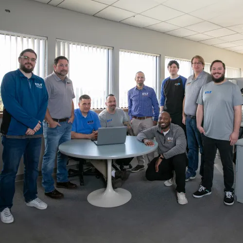 Group of nine diverse colleagues in casual attire posing in a bright office around a round table with a laptop.