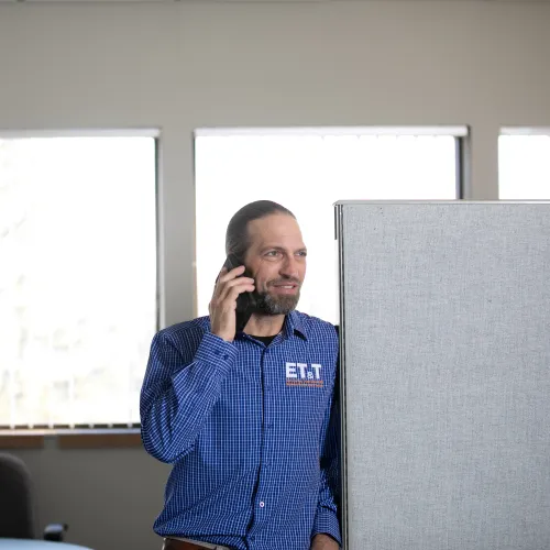 Man in blue checkered shirt talking on phone in office cubicle with bright windows behind.
