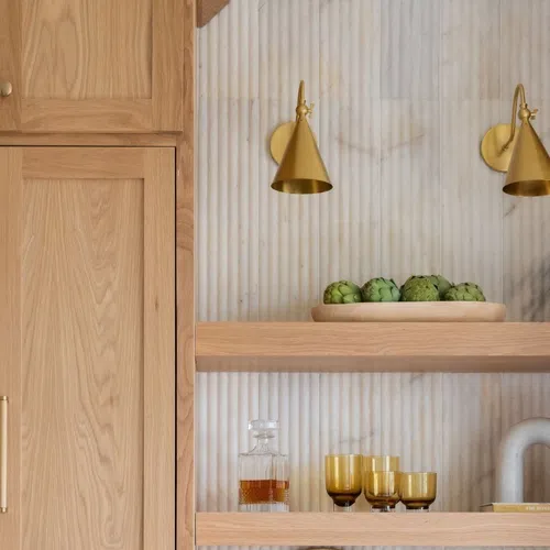 Modern wooden kitchen shelves with artichokes, amber glassware, and golden wall sconces on marble wall.